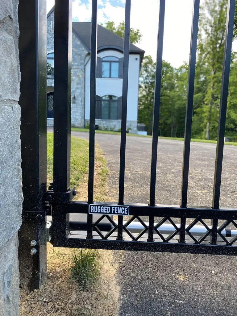 Decorative automatic metal gate close up with the Rugged Fence logo attached to it with a house in the background