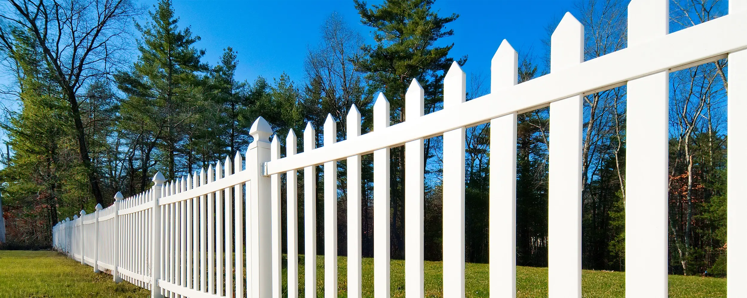 White Picket fence at an angle with trees behind it and green grass below it.