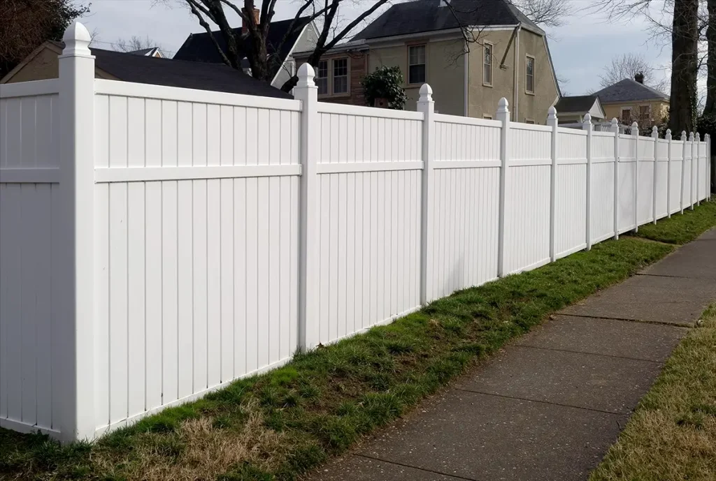 A tall white vinyl privacy fence runs alongside a sidewalk and grassy strip in a suburban neighborhood, separating the yard from the public walkway.